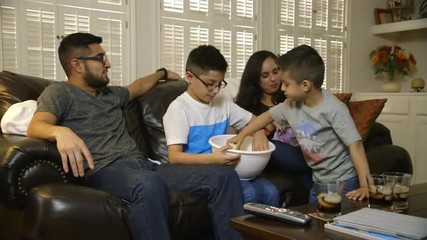 An attractive Hispanic family with two cute boys enjoying popcorn as they all watch TV together.
