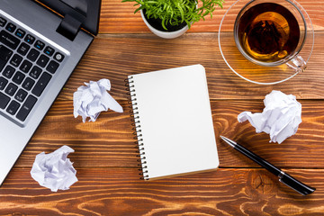 Office table desk with supplies, white blank note pad, cup, pen, pc, crumpled paper, flower on wooden background. Top view