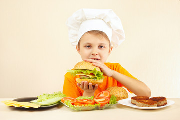 Young funny boy in chefs hat enjoys a cooking tasty hamburger