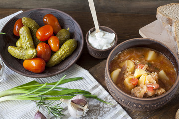 Traditional ukrainian vegetable soup - borsch, marinated tomatoes and cucumbers, sour cream, sliced bread, herbs and garlic at dark wooden table.