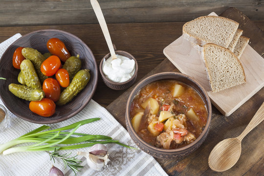 Traditional ukrainian vegetable soup - borsch, marinated tomatoes and cucumbers, sour cream, sliced bread, herbs and garlic at dark wooden table.