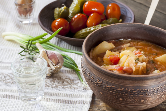 Traditional ukrainian vegetable soup - borsch, marinated tomatoes and cucumbers, sour cream, sliced bread, herbs and garlic, vodka in glass cup at dark wooden table.