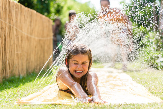 Wasser Spaß im Garten - Mädchen mit Rasensprenger kühlt auf Wasserrutsche im Sommer ab