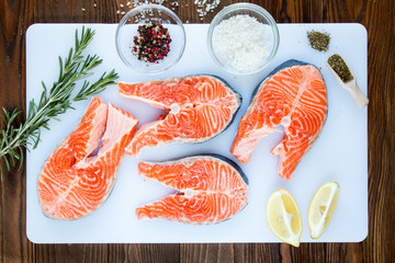 steaks of salmon with a sprigs of rosemary, lemon, pepper,  sea salt, , herbs in wooden scoop on a white cutting board on wooden background, top view.