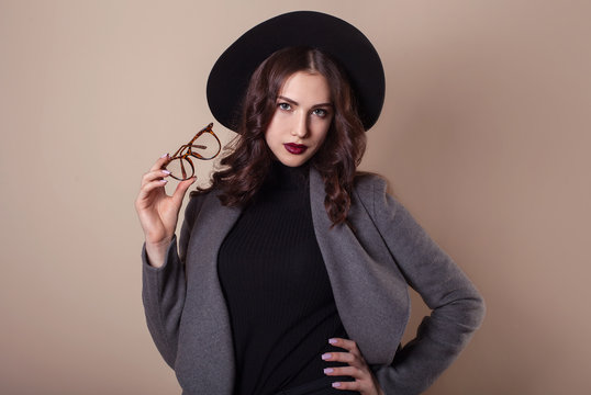 Fashion portrait of young pretty hipster brunette model woman in hat glasses and warm coat. Beige background copy space
