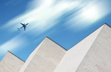 Flugzeug fliegt über modernes weißes Backsteingebäude mit Bewegungsunschärfewolken im Hintergrund © BOOCYS