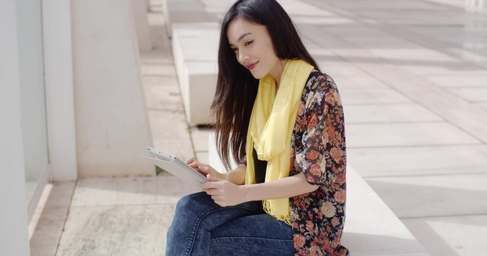 Young woman sitting enjoying the sunshine reading her tablet on a bench in an urban mall or walkway with a smile of pleasure