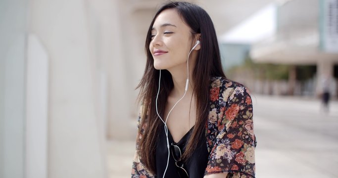 Trendy young woman listening to music on her earphones in town sitting cross legged on a bench in a pedestrian walkway with a smile of pleasure.