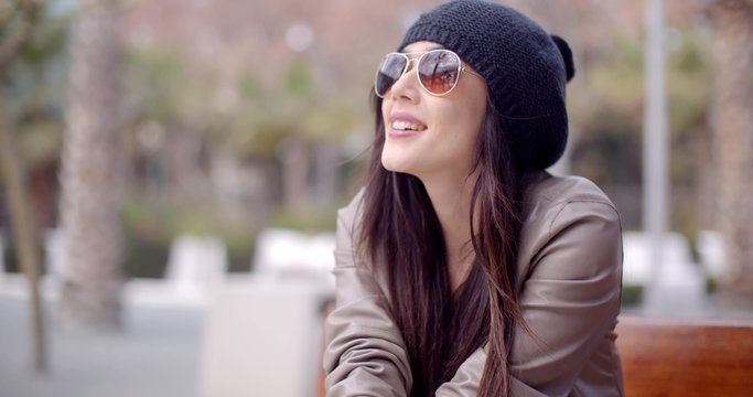 Gorgeous young woman wearing sunglasses and a woolly cap sitting daydreaming on a wooden bench outdoors looking up with a happy smile