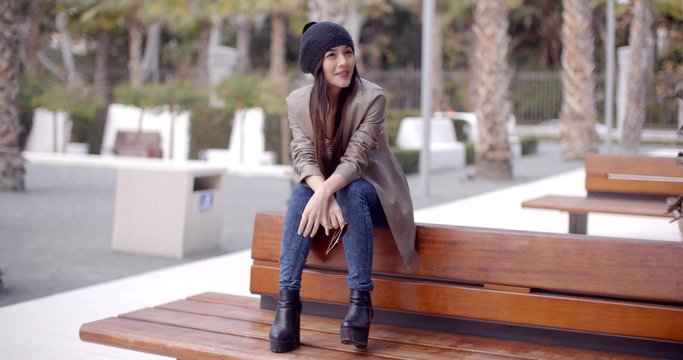 Fashionable young woman in a trendy modern outfit and boots sitting on an outdoor wooden bench in a park waiting for someone with a look of expectation.