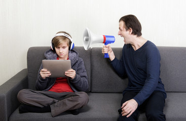 Tween son in headphones looks at the digital tablet display while his father yells at him through a megaphone