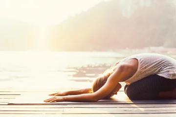 Rolgordijnen Zonnegroet yoga. Jonge vrouw doet yoga aan het meer, badend in het zonlicht. © Microgen