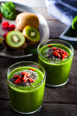 Fresh green smoothie sprinkled with chia seeds and goji berries in the background fruits and vegetables on wooden table.