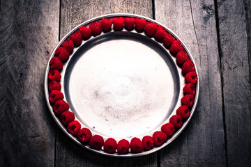 Raspberries arranged around the edge of the old metal plate on wooden background top view.