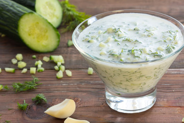 Tzatziki – traditional greek sauce in glass bowl and ingredients – cucumber, garlic, dill on wooden dark background