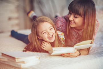 Portrait of a smiling young cute mother and daughter reading a book lying and relax in the bed in a...