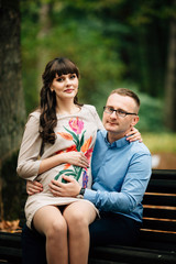 Beautiful and happy pregnant couple relaxing outside in the autumn park sitting on bench.