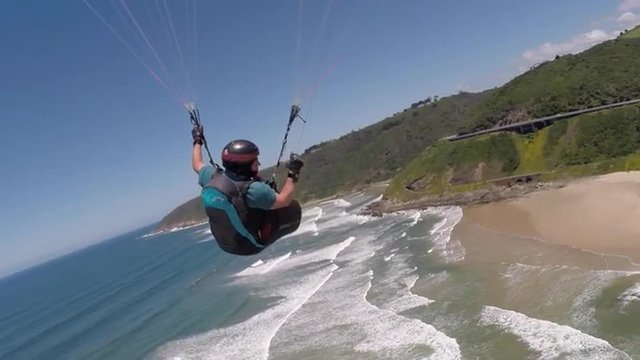 POV Paraglider Pilot Chase Cam following behind pilot making a 360 degree turn over the beach in Wilderness, South Africa with spectacular view of the coastline.