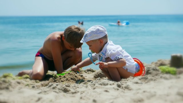 Sunny summer weather, vacationers - children, teen boy and toddler baby  playing with toys in the sand on the beach  and sunbathing, wave  on a sandy beach and above them the beautiful sky 