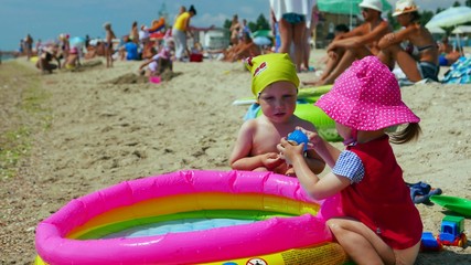 Sunny summer weather, two toddler vacationers  - children, little girl and toddler baby boy playing with toys in inflatable paddling pool on the beach  and sunbathing. Childhood dreams and memories.