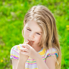 Adorable blond little girl with long hair eating ice-cream on summer sunny day, outdoor portrait on green grass background