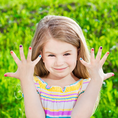 Cute smiling blond little girl with many-coloured manicure, outdoor portrait on green grass background