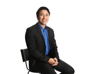 Man in his 20s sitting down on a chair against a white background, wearing a black suit with a dark blue shirt. Looking at camera with a big toothy smile.