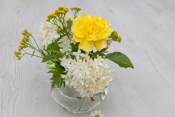 Bright summer bouquet of white hydrangea, yellow roses and tansy flowers  in a glass vase on a white wooden table