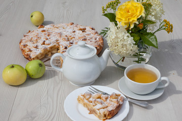 Apple pie and camomile tea set on the white wooden table decorated with fresh flowers and apples.
