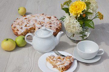 Apple pie and camomile tea set on the white wooden table decorated with fresh flowers and apples.