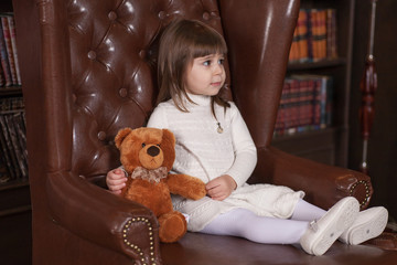An adorable 2-year-old girl is sad. Little Princess and her toy bear sitting in a chair. Portrait in full growth in the interior