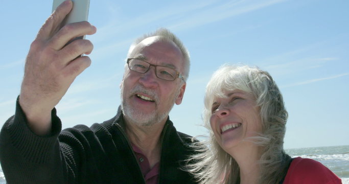 A smiling senior couple take a selfie photo of themselves on the beach. Shot in 4K UHD slow motion.