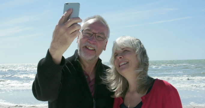A smiling senior couple take a selfie photo of themselves on the beach. Shot in 4K UHD slow motion.