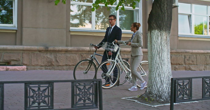 Tracking shot of businessman and businesswoman in suits walking with their bicycles down street and talking on the move