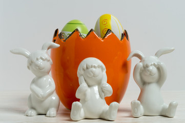 Three white ceramic rabbits with egg shell helmets in front of a egg shaped flowerpot filled with hand painted easter eggs
