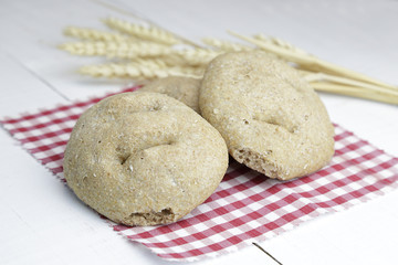 Three pieces of brown bread and a branch of wheat over a red and white checkered napkin on a white wooden table