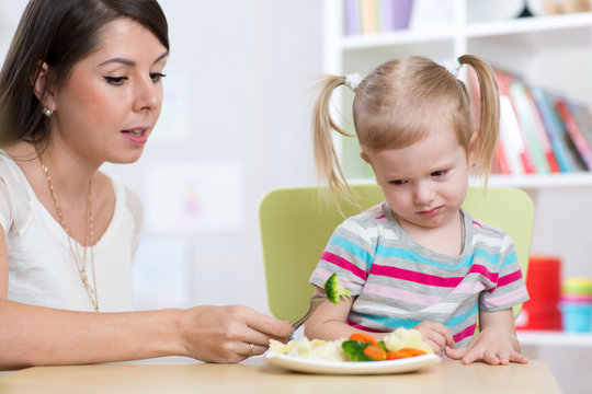 Child Girl Looks With Disgust At Healthy Vegetables. Mother Convinces Her Daughter To Eat Food.