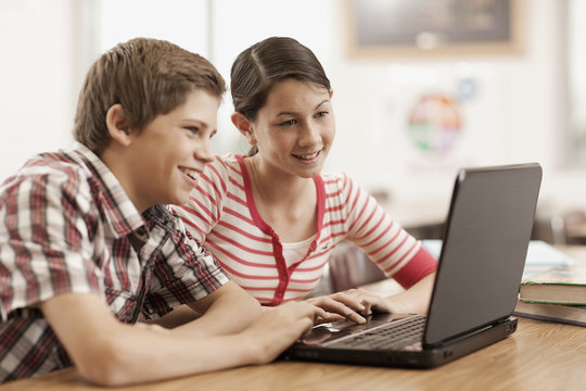 Two children, a boy and girl sitting at a desk, using a laptop computer. 