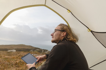 A man sitting in the shelter of a tent looking out, holding a digital tablet. 
