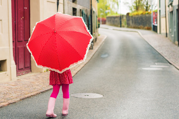 Cute little girl walking down the street, wearing pink rain boots, holding polka dot red umbrella, back view