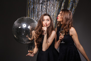 Two amusing women showing silence gesture and drinking champagne - obrazy, fototapety, plakaty