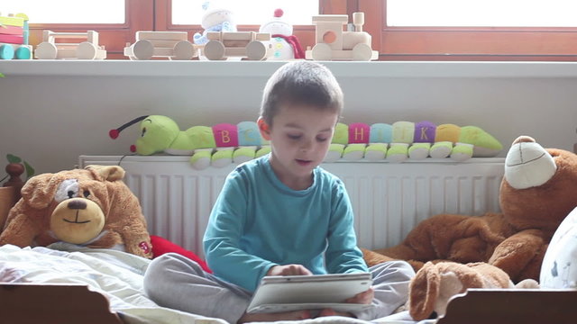 Cute little boy,sitting in bed in kids room, playing on tablet happily, stuffed toys around him