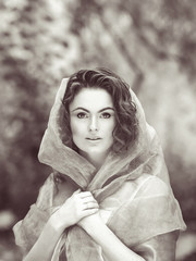 Black and white close-up beauty portrait of young white Caucasian girl with wavy curly hair covered with hood clothes gauze, in park outside on summer day