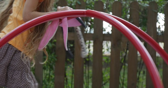 Girl is Swinging on The Carousel Holding Pink Toy Tilda Rabbit Little Girl With Long Fair Hairs in Orange T-Shirt is Playing at Playground Wooden Fence