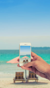 Männliche Hand haltet ein weißes Smartphone und fotografiert damit einen idyllischen Strand mit Sonnenliegen