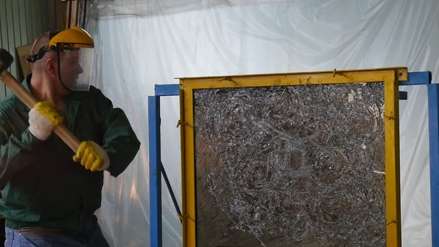 Worker In Uniform And Protective Screen, Yellow Gloves, is Beating the Glass Sheet by Hummer, Testing of Bulletproof Glass, Tempered Glass Breaking