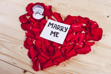 Heart by red roses petals on wooden board with a notes Marry me and proposal ring
