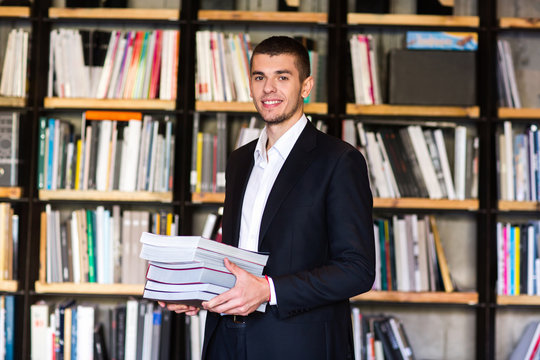 Student in library. Handsome young man holding books and smiling while standing in library