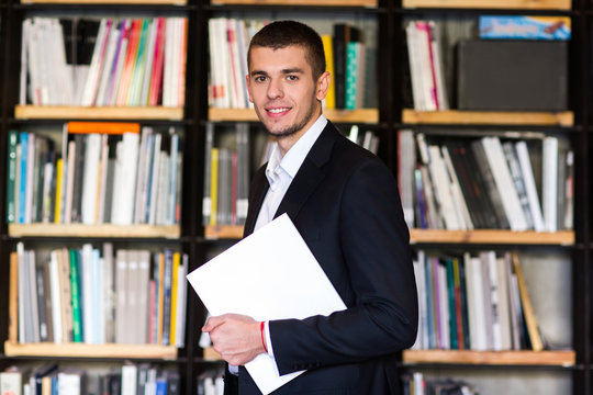 Student in library. Handsome young man holding books and smiling while standing in library