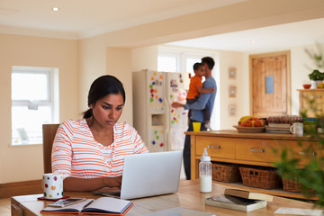 Family In Kitchen With Mother Using Laptop At Table